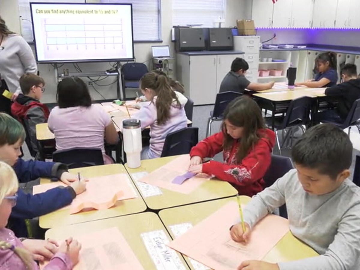 Third and fourth graders at their desks folding paper strips into equal parts, teacher at the board